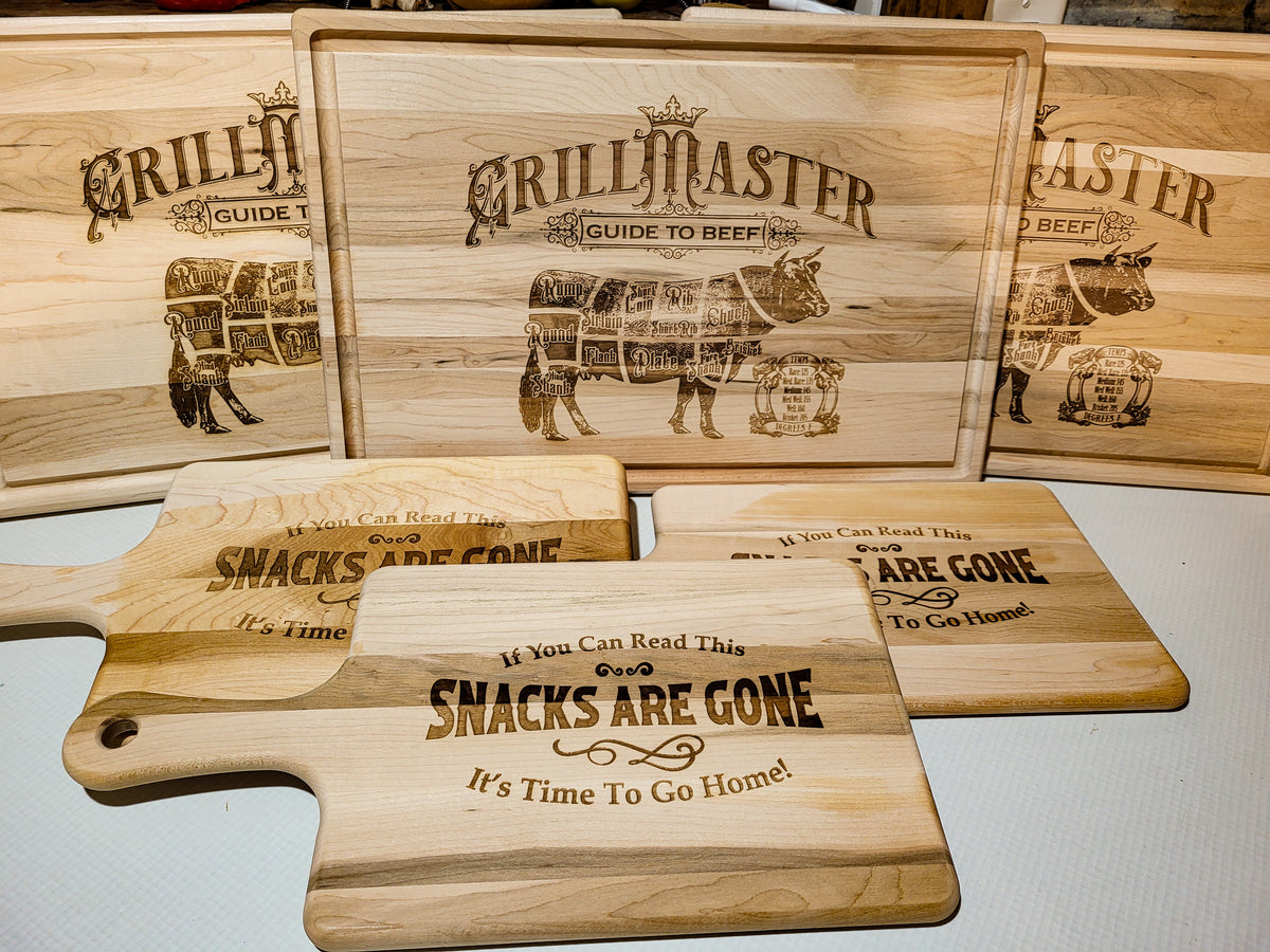 A set of wooden cutting boards and serving boards with engraved text and illustrations. The center board reads 'GRILLMASTER GUIDE TO BEEF' with a beef carcass graphic, while the foreground boards read 'If You Can Read This Snacks Are Gone It's Time To Go Home!' and 'GRILLMASTER GUIDE TO SNACKS'.