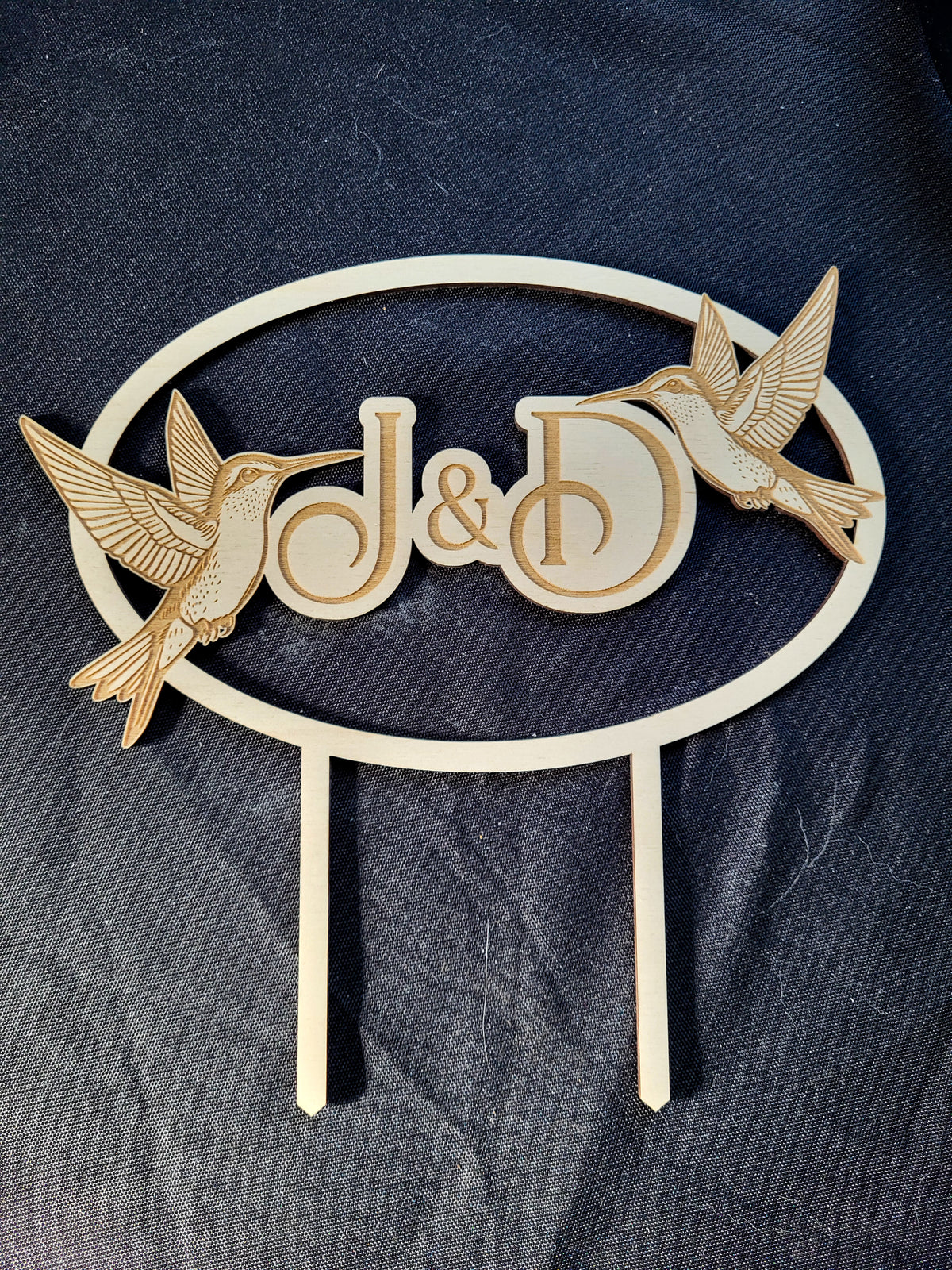 A wooden custom garden stake featuring an oval frame with the initials 'J&D' in the center and two laser-cut hummingbirds on either side, displayed on a dark textured fabric background.