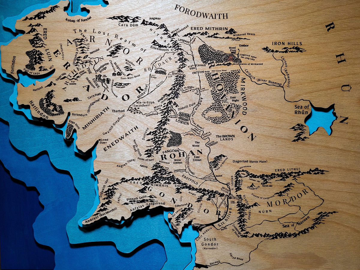 A 3D wooden map of Middle Earth from Lord of the Rings, featuring carved terrain details, black engraved text, and layered blue wood coastlines, displayed against a dark wood plank background.