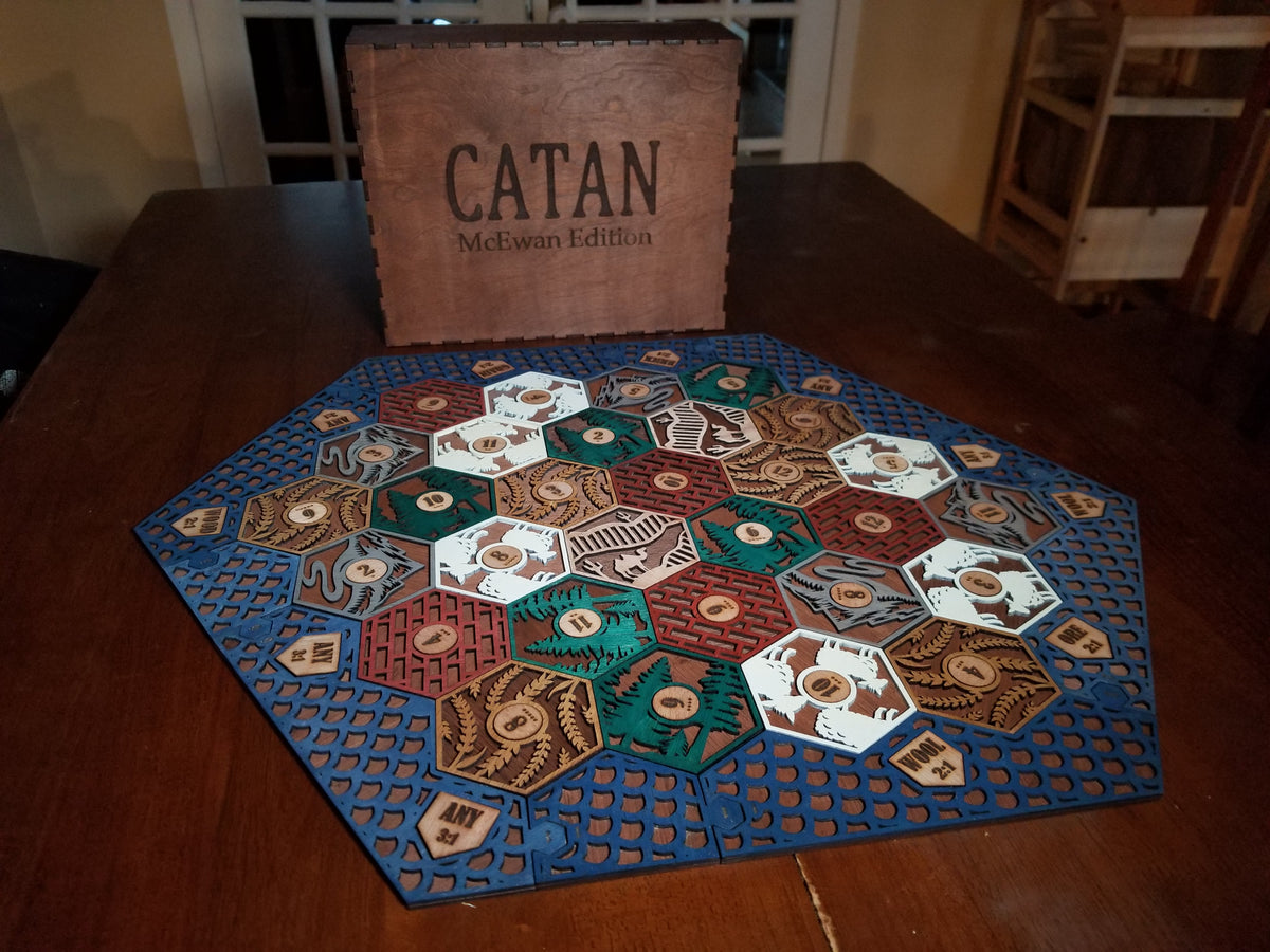 Custom Catan board game set on a wooden table with its wooden box behind it. The board features a blue border and multiple colored hexagonal tiles representing resources and buildings.