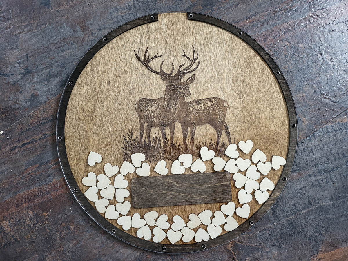 A round wooden plaque featuring an engraved stag and deer design, bordered by a dark metal rim and decorated with small white heart-shaped wooden tokens arranged around a blank central plaque, set against a rustic dark wood background.