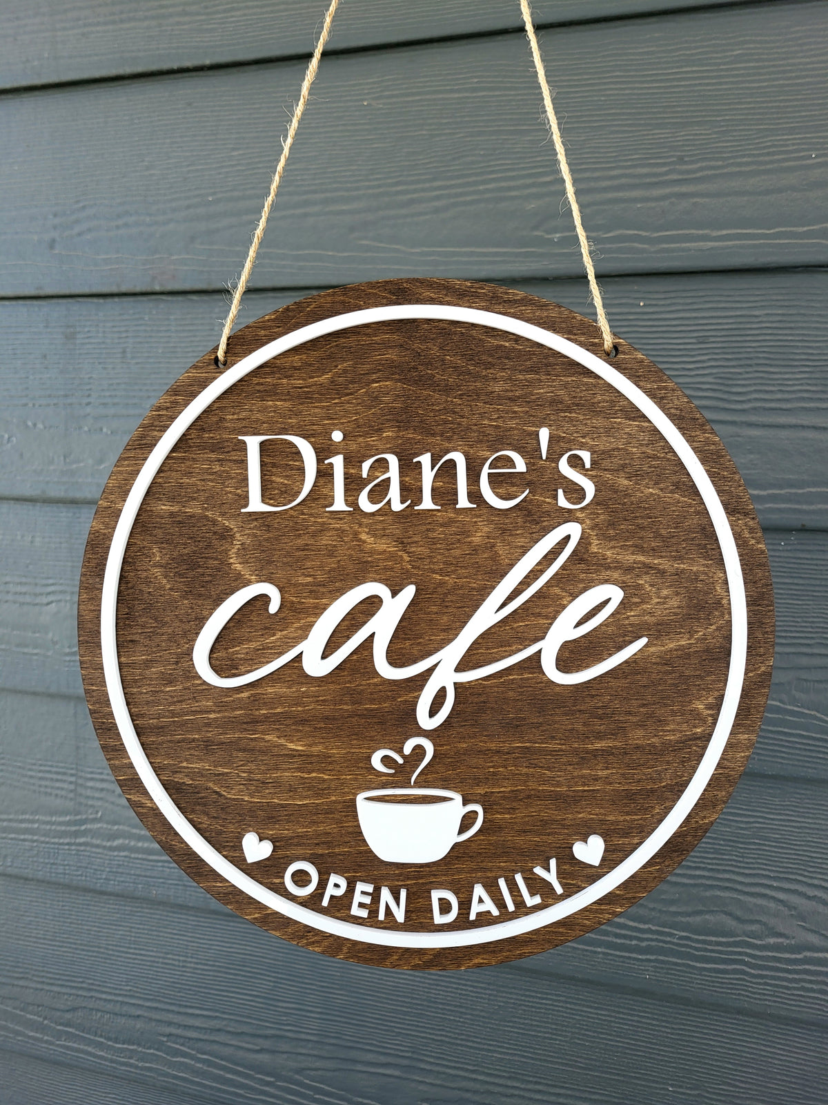 A round wooden hanging sign for 'Diane's Cafe' with white text and a coffee cup graphic, suspended by twine against a dark gray wood-paneled background.