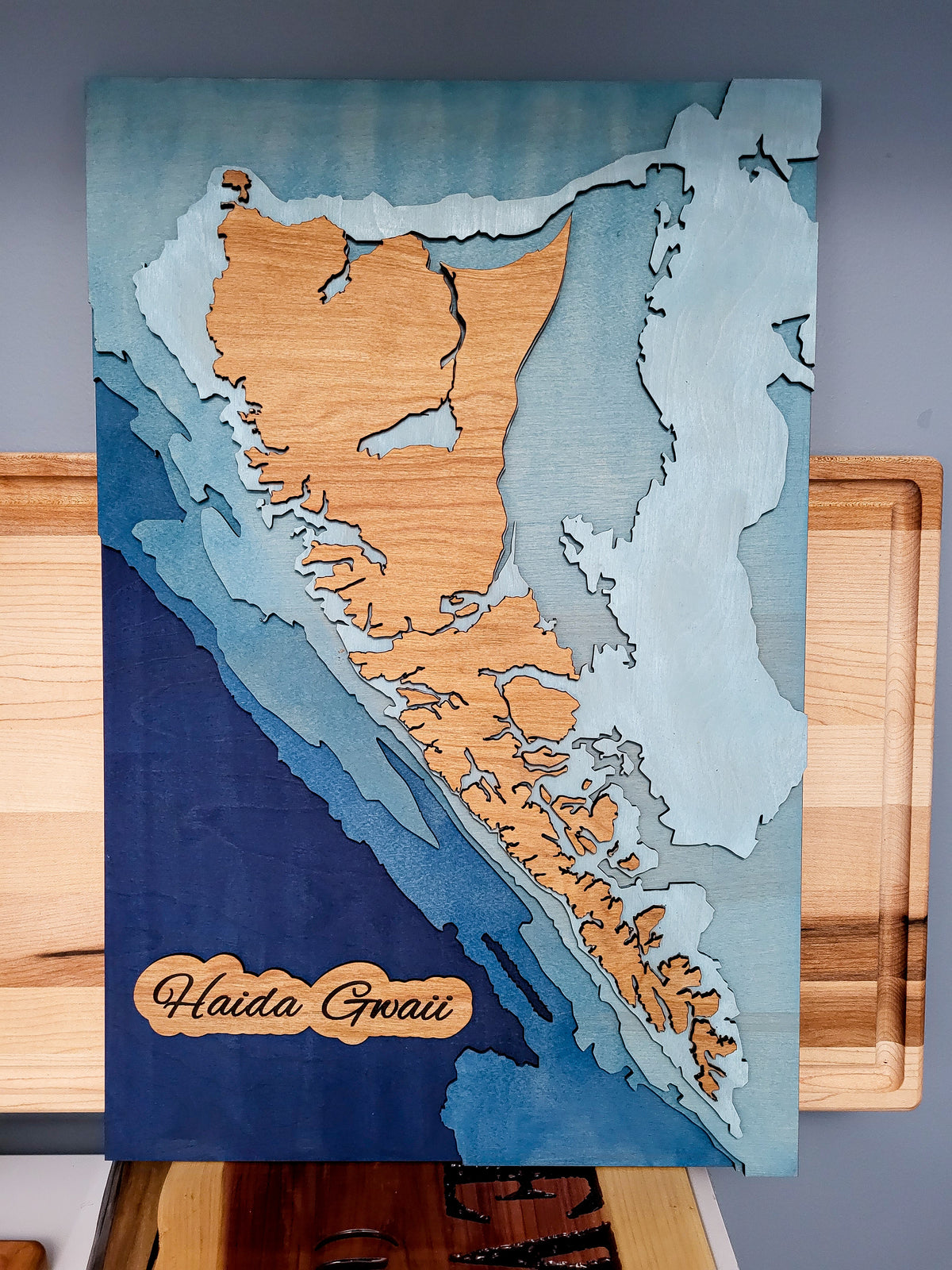 A 3D layered wooden map wall art piece of Haida Gwaii, featuring natural wood grain layers over blue and gray watercolor-style backgrounds, displayed on a wooden stand.