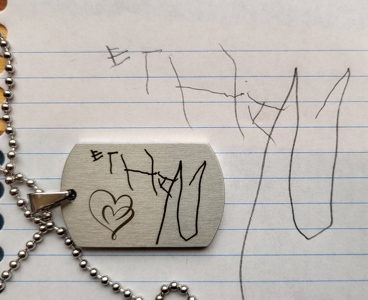 A silver metal dog tag necklace with engraved black markings, including a heart and stylized text, resting on blue-lined notebook paper with handwritten marks.
