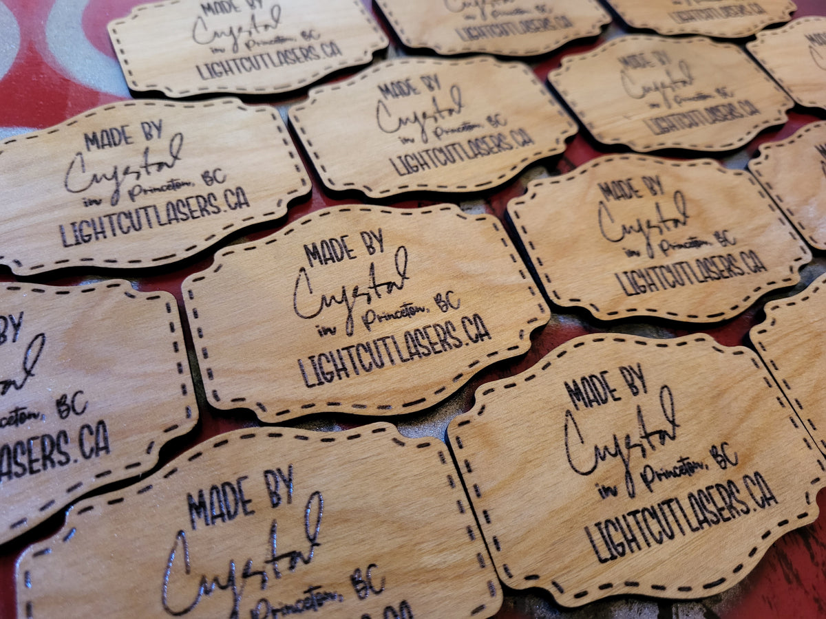 Multiple laser-engraved wooden tags with stitched border designs, featuring the text 'MADE BY Crystal in Princeton, BC LIGHTCUTLASERS.CA', arranged on a red patterned surface.