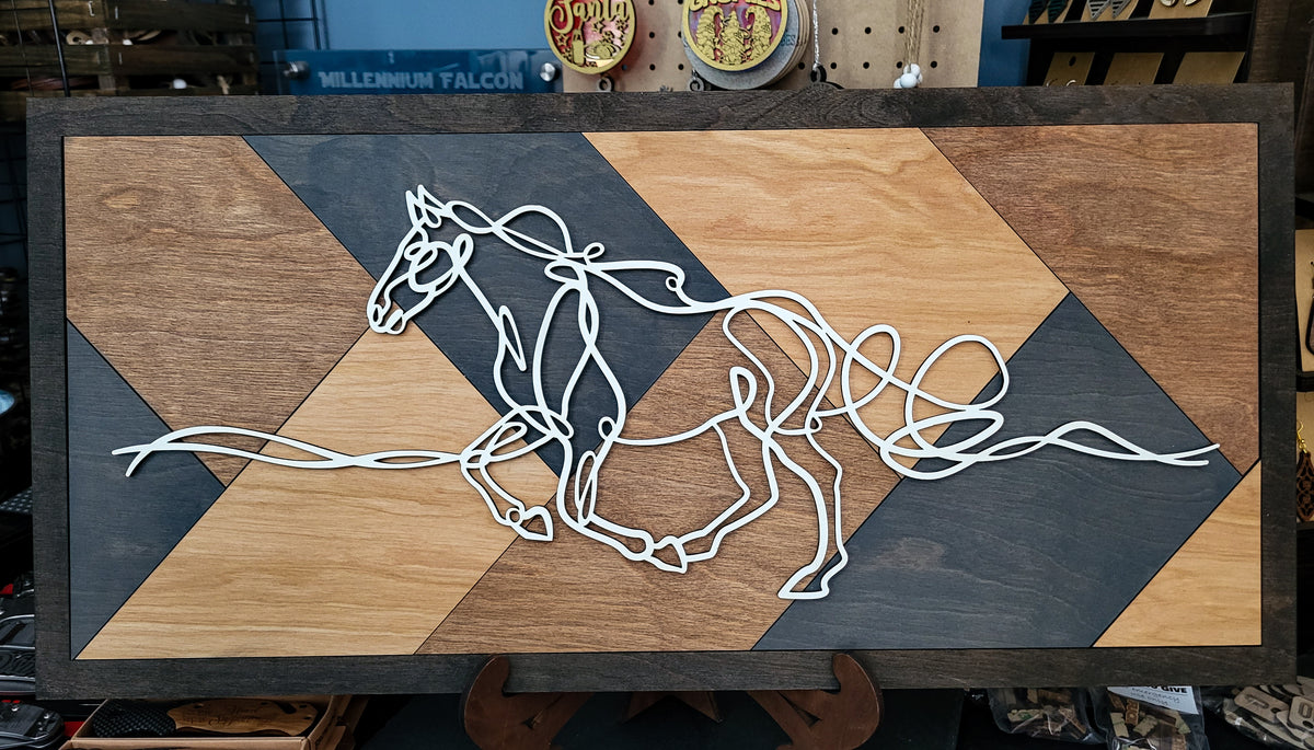 A rectangular wooden wall art piece featuring a stylized white wire horse running across a geometric background of light and dark wood panels, displayed on a stand in a shop setting.