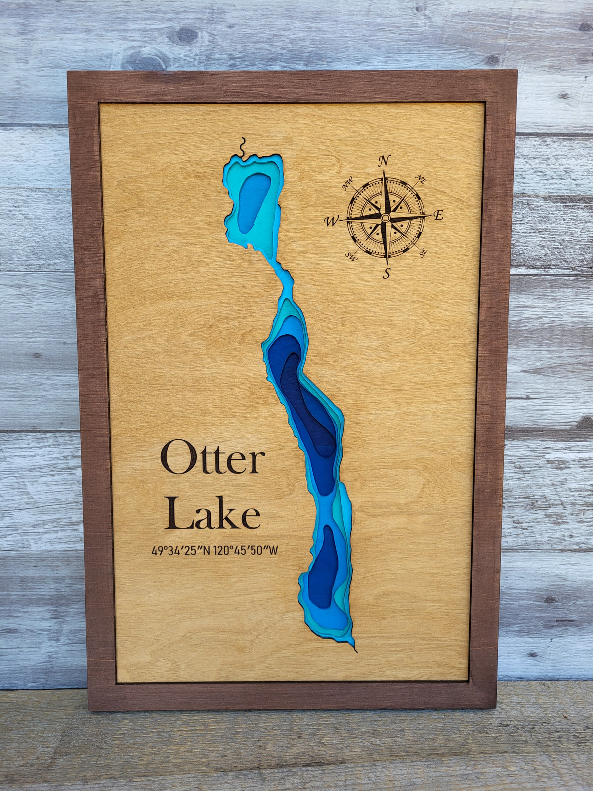 A framed wooden Otter Lake map print with layered blue and teal depth contours, a compass rose, and location text, displayed against a rustic gray wood plank background.