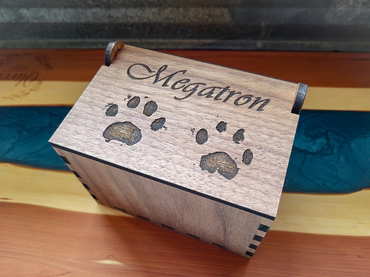 A small wooden keepsake box engraved with the name 'Megatron' and paw prints, resting on a wooden surface with a blue inlay.