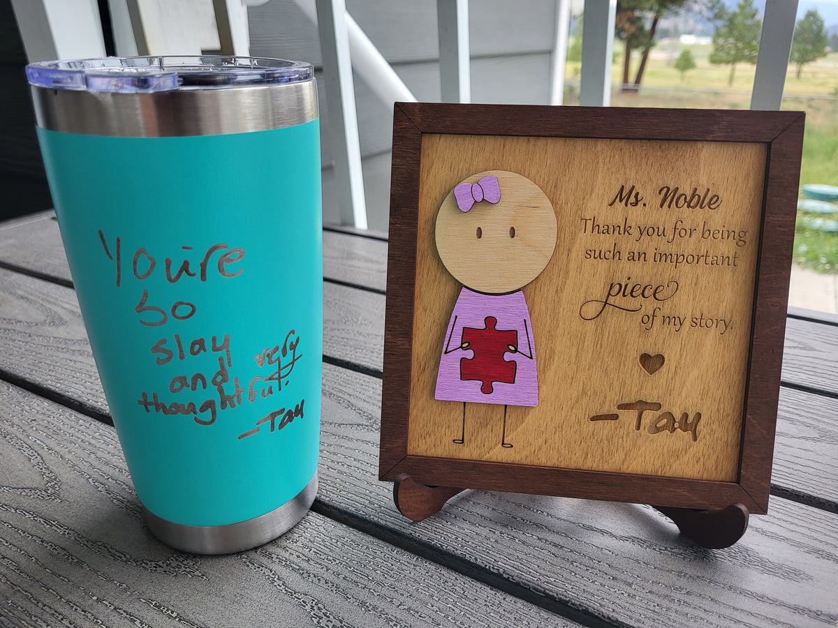 A turquoise insulated tumbler with a stainless steel rim and lid sits beside a small wooden engraved plaque on a gray deck railing. The plaque features a cartoon girl with a purple bow and pink puzzle-piece dress