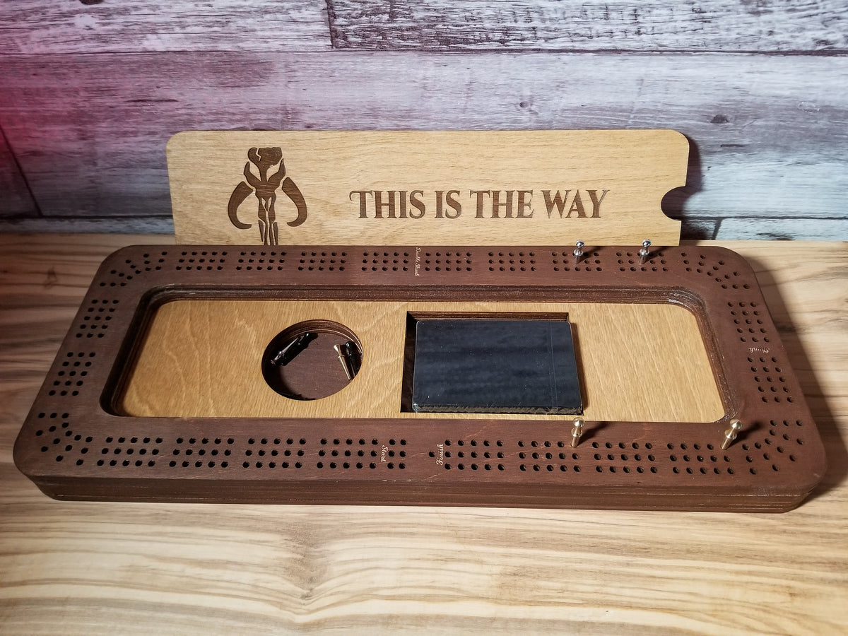 A wooden gaming board set with a dark brown perforated board, a light wood playmat, a black rectangular accessory, and an engraved backplate that reads 'THIS IS THE WAY' with a mandalorian, displayed on a wooden surface.